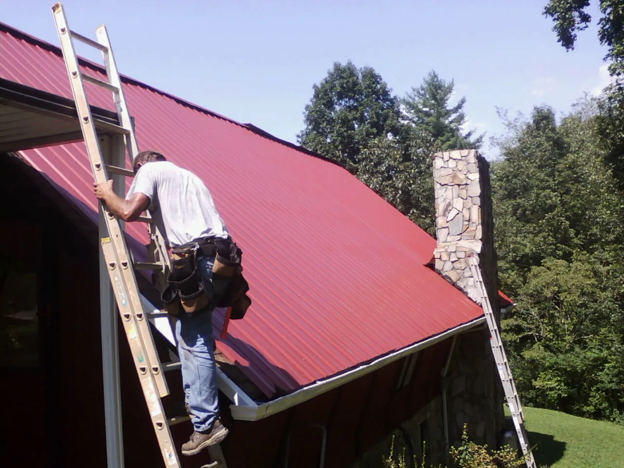 Roofer on a ladder beside a red metal roof and stone chimney — Soffit & Fascia Repair by Roofing Brothers in Pownal